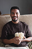 Fröhlicher, lockig behaarter Afroamerikaner in Freizeitkleidung, der sich auf einem bequemen Sofa ausruht, in die Kamera schaut und leckeres Popcorn isst, während er am Wochenende Fernsehkanäle schaut
