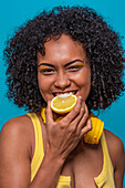 Portrait of content African American female model with curly hair smiling and looking at camera while biting juicy lemon against blue background