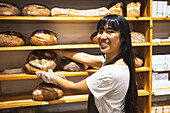 Side view of cheerful Asian female seller smiling and looking at camera while standing near shelves with assorted bread loaves of bakery