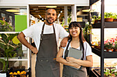 Positive young African American sales staff in apron smiling and looking at camera while male standing with hand on shoulder of female with tablet