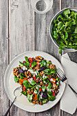 Top view of appetizing fresh salad with green leaves and walnuts seasoned with sauce and served on white plate with spoon and fork near bowl with spinach