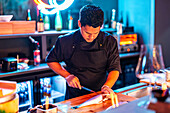 Focused male chef wearing gloves and black uniform slicing fish with knife on illuminated wooden counter at modern sushi bar