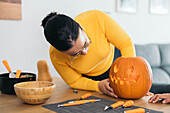 Focused young Hispanic female looking down while standing with anonymous friend with fingers on table and putting Jack o Lantern in Halloween pumpkin