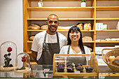 Positive young multiracial coworkers in aprons smiling and looking at camera while standing behind counter of bakery shop during work time