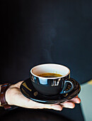Crop anonymous person showing cup of hot aromatic coffee with saucer on black background indoors