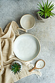 Draufsicht auf Keramikgeschirr, bestehend aus Schüssel, Teller und Holzlöffel, neben Serviette und Topfpflanzen auf grauer Fläche