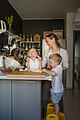 Caucasian young mother smiling and looking at twin sons eating banana and boy cutting fruit on chopping board while cooking together in kitchen