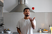 Confident young ethnic African American male in white t shirt throwing up red tomato and listening to music in true wireless earbuds while preparing healthy food in kitchen