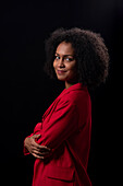 Side view of smiling young Hispanic female model with Afro hairstyle in red coat standing arms crossed against black background