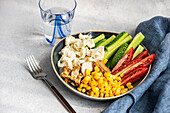 Schüssel mit fünf Zutaten, Blumenkohl, Tomate, Gurke, gekochtem Mais und gegrilltem Hühnerfleisch auf grauer Fläche, neben Serviette, Glas und Gabel