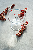 A clear glass cake stand on a textured surface, adorned with small red berries scattered around it