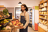 Side view of positive young bearded ethnic male seller with long curly hair in apron smiling and looking at camera while demonstrating box with fresh ripe peaches in supermarket
