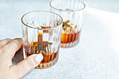 Closeup of crop hand of anonymous woman touching glass of whiskey with ice and orange peel placed on gray background