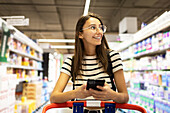 Positive young female customer in casual clothes and eyeglasses using smartphone while shopping alone at supermarket