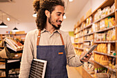 Positive bearded man in apron smiling and looking at screen while browsing smartphone and standing near shelves with various products in grocery shop
