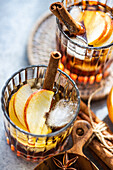 Fresh apple cider cocktails garnished with cinnamon sticks, star anise, and apple slices in a crystal glass filled with ice cubes on a textured background