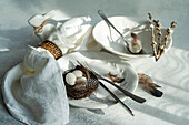 Closeup of elegant Easter dining arrangement, featuring pristine white plates with a delicate embossed design with woven nest containing speckled eggs rests atop one plate, accompanied by fork and knife, feathers and branches