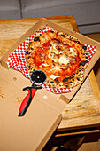 Blick von oben auf eine appetitliche gebackene Pizza mit Tomaten und Käse mit knuspriger Kruste, die in einem Karton mit Messer auf dem Tisch liegt