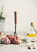 Rohes Fleischsteak mit Zutaten und Fleischgabel auf Küchentisch mit Rosmarin, Pfeffer und Olivenöl. Kochen zu Hause mit frischem Fleisch. Vorderansicht. Stilleben