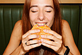 Closeup of young woman with red hair and piercing with closed eyes sitting at table and enjoy eating hamburger in restaurant