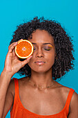 Serious young Hispanic female with curly hair covering eye with slice of orange and with closed eyes against blue background