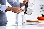 Crop anonymous adult male chef in apron standing in kitchen and pouring ingredients from saucepan into glass during breakfast preparation in modern kitchen in daylight