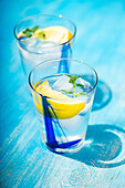 High angle of Summer cocktails with lemon vodka, slices of lemon and wild mint leaves served with ice placed on blue table