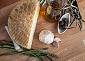 A wedge of artisan cheese is presented with olive oil, garlic, asparagus, and olives on a wooden background.