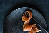 Top view of closeup of healthy salmon slice served on black plate near fork against dark surface