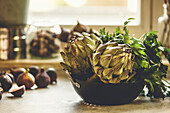 Whole artichokes in bowl with parsley on kitchen table with figs at window background with natural light. Healthy Mediterranean food cooking. Front view.