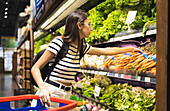 Side view of young female customer in casual clothes with shopping basket choosing fresh carrots while standing near shelves in supermarket