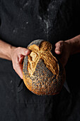 A pair of hands holding a freshly baked artisan bread loaf adorned with a poppy seed crust, against a dark background