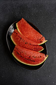 Top view of slices of watermelon served on black plate against dark surface