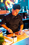 Focused Male chef wearing gloves and black uniform with concentration slicing meat with knife on counter in kitchen at modern illuminated sushi bar