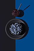 Frische Blaubeeren in einer schwarzen Schale mit einer Tasse Blaubeermarmelade auf einem geteilten blauen und schwarzen Hintergrund, der ein Spiel von Licht und Schatten zeigt