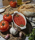 Reife Tomaten und ungeschälter Knoblauch auf einem Tisch neben einer Schüssel mit zerdrückten Tomaten, Vollkornbrot auf einer Serviette, einem Schneidebrett und Öl