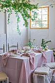 Elegant table served with plates and flowers near yummy cake on pink tablecloth placed near bench covered with cloth against potted plant and white brick wall