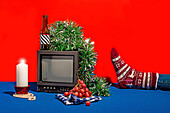 Vintage-Fernsehgerät, umgeben von einer Reihe von Objekten, darunter eine Flasche mit gestreiftem Etikett, frische Trauben auf einem karierten Tuch, eine weiße Kerze und grünes Lametta, alles vor einem roten Hintergrund auf einem blauen Tisch mit einem Gertenfuß mit festlich verzierter Socke