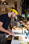 Side view of bearded male cook in apron standing and adding cream cheese on seaweed wrap placed on bamboo roll while looking down and working in modern kitchen