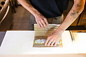 From above of unrecognizable tattooed male chef in apron standing near table and making sushi roll with bamboo mat with hands while working in kitchen