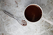 Top view of melted hot milk chocolate in metal saucepan with whisk placed on marble counter for pastry cooking