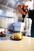 Anonymous male chef in black shirt standing at table in kitchen and blowing gas flame on delicious uramaki sushi while preparing to serve food against blurred interior