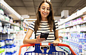 Positive young female customer in casual clothes and eyeglasses using smartphone while shopping alone at supermarket