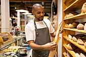 Positive young African American male seller in apron putting fresh baked bread into paper bag while standing near shelves during work process