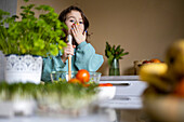 Porträt eines süßen, überraschten kaukasischen Mädchens mit bunten Fingernägeln, das den Mund mit der Hand bedeckt und einen Salat in der Küche zu Hause zubereitet