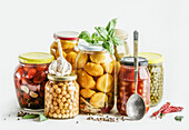 Preserved vegetables selection in glass jars with herbs, spices and spoon at white background: conserved chickpeas, zucchini, beans and tomatoes. Healthy sustainable cooking. Front view.