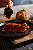 High angle of crop anonymous cook pouring delicious sauce on grilled pork ribs served on plate garnished with baked potatoes