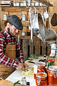 Side view of mature focused chef in apron and beret looking down while chopping onions, surrounded by a rustic setting with hanging kitchenware and jars of homemade preserves