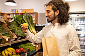 Portrait of young ethnic man with curly hair in casual clothes standing near shelves with assorted fresh vegetables and fruits while shopping indoors