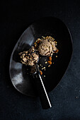 Draufsicht auf einen schwarzen Keramikteller mit einer Kugel Eis, der neben einer Kugel Schokoladeneis mit Erdnüssen und Walnüssen steht
